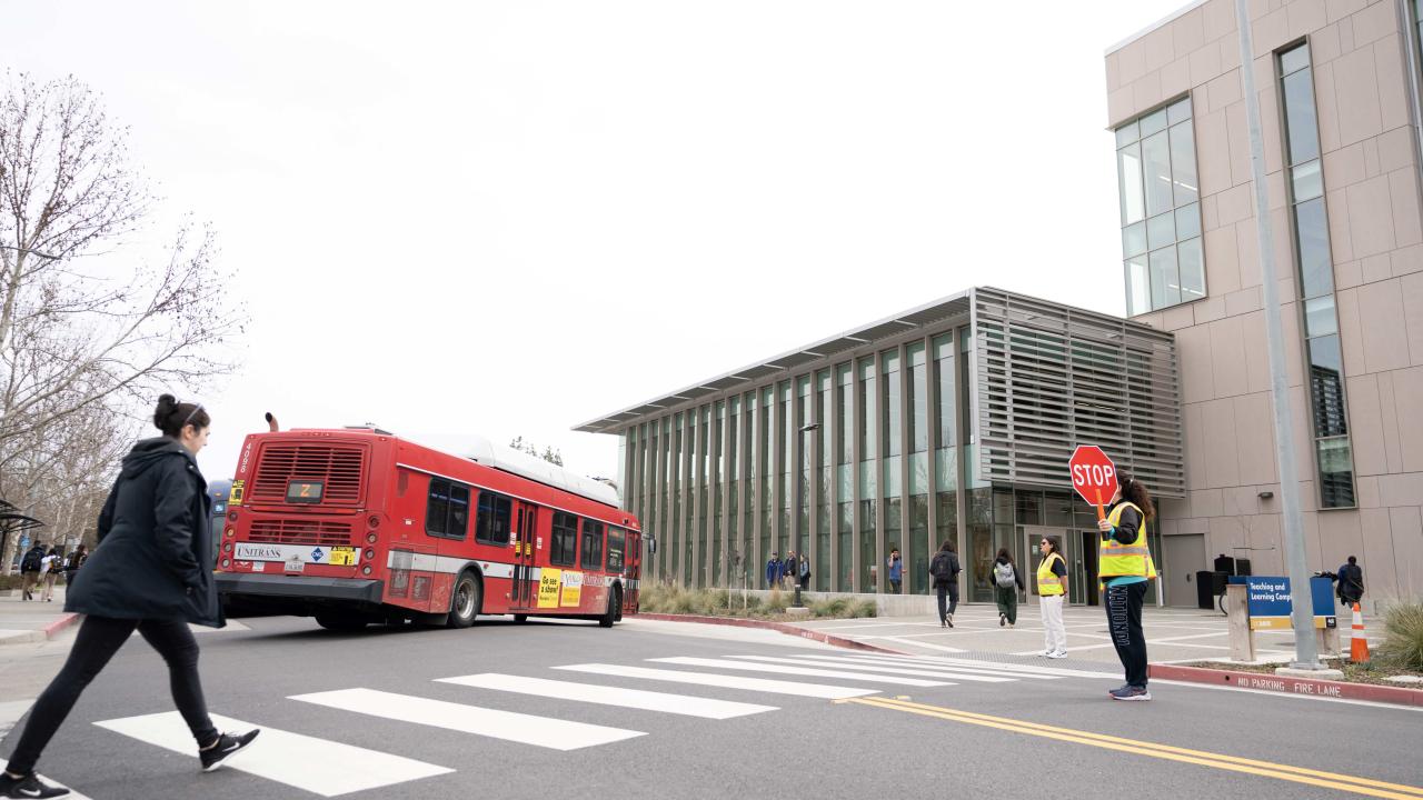 Pedestrian walks across crosswalk, while traffic ambassador holds a stop sign, in front of UC Davis Teaching and Learning Complex