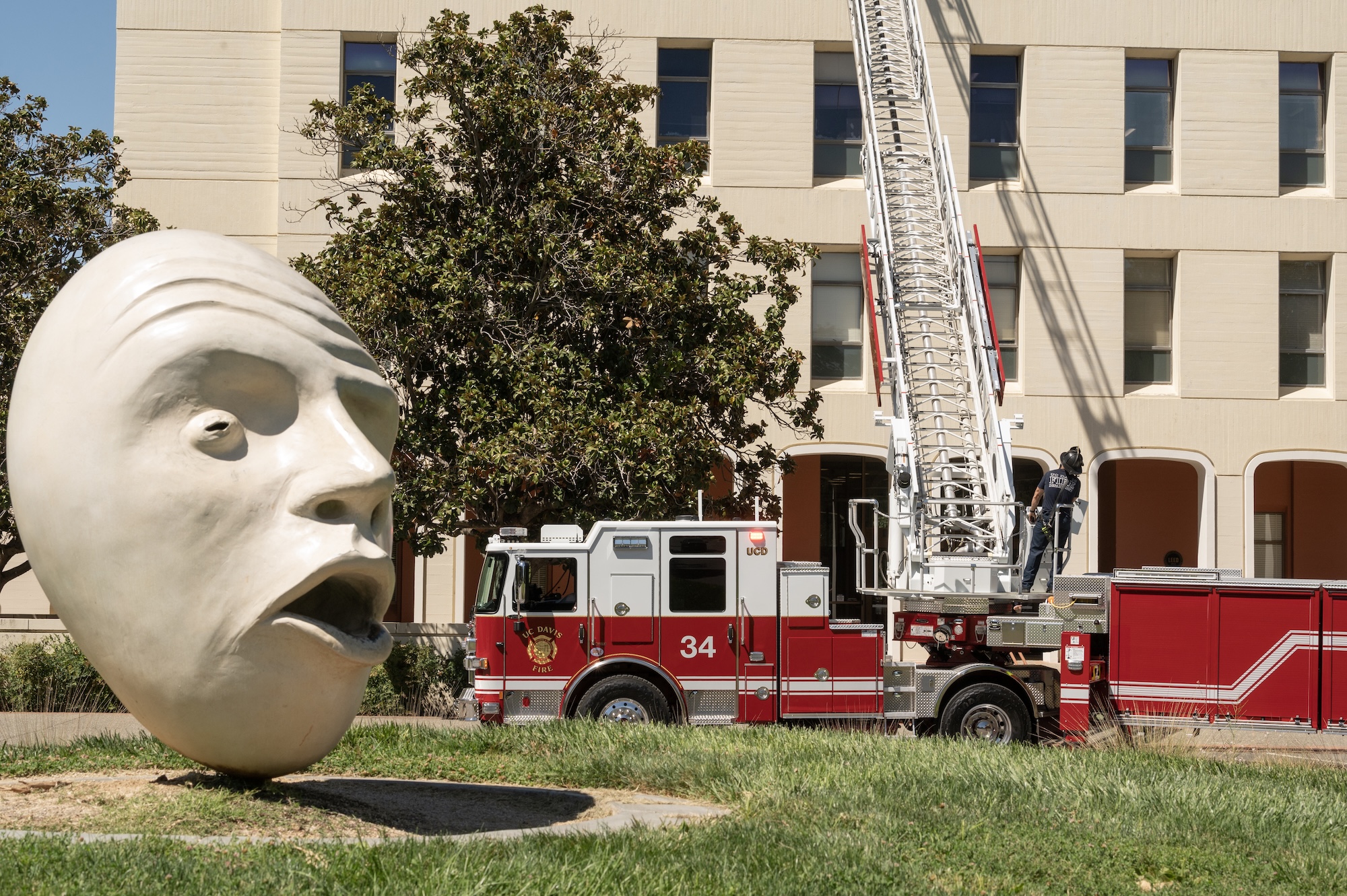 Exterior shot of Mrak Hall. In the foreground is one of the "See No Evil/Hear No Evil" Egghead statues. In the background, a firetruck has its ladder extended toward Mrak Hall and a firefighter stands on top of the fire engine, at the base of the ladder.