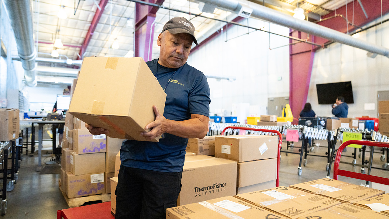 A UC Davis Distribution Services staff member moving a cardboard box onto a pallet that has several other boxes on it.