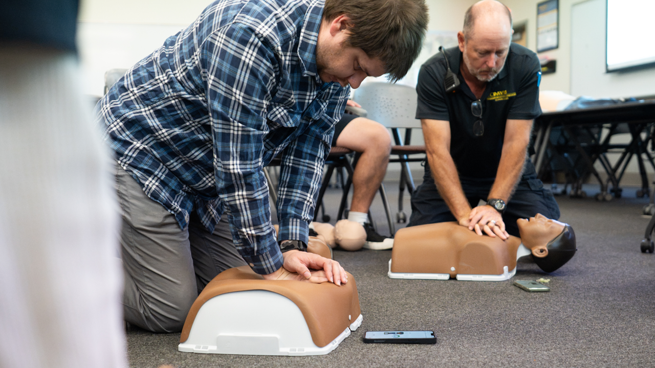 Two men kneeling in a classroom practicing CPR on adult training mannequins