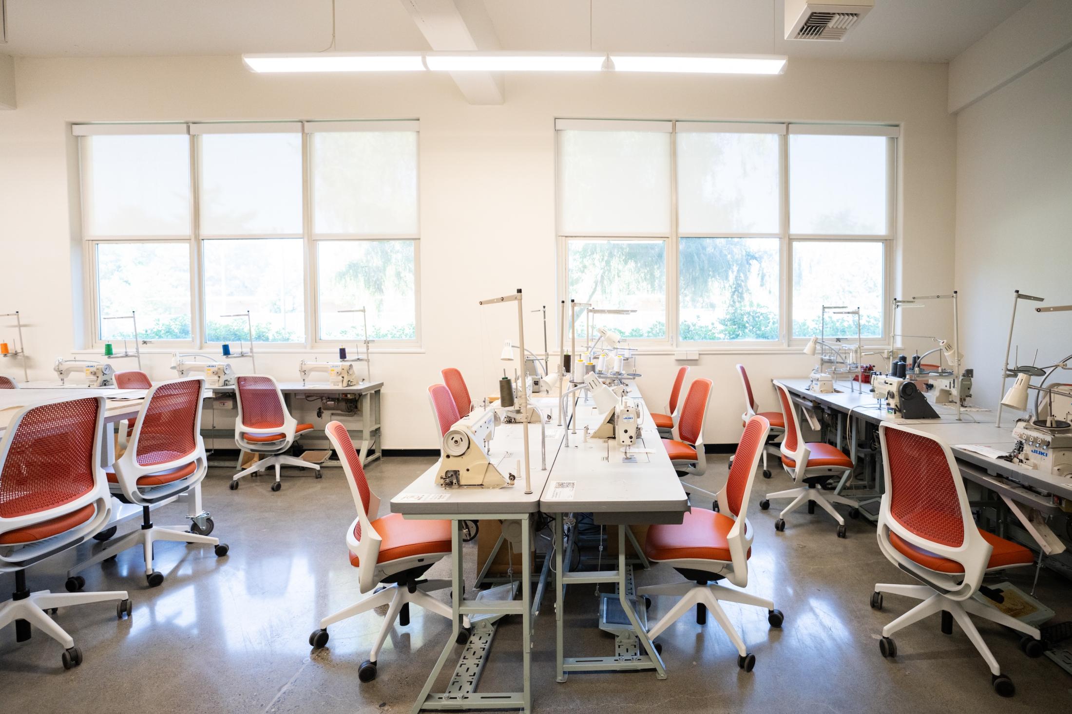 Sewing lab with sewing machines, rows of worktables, orange chairs and large windows
