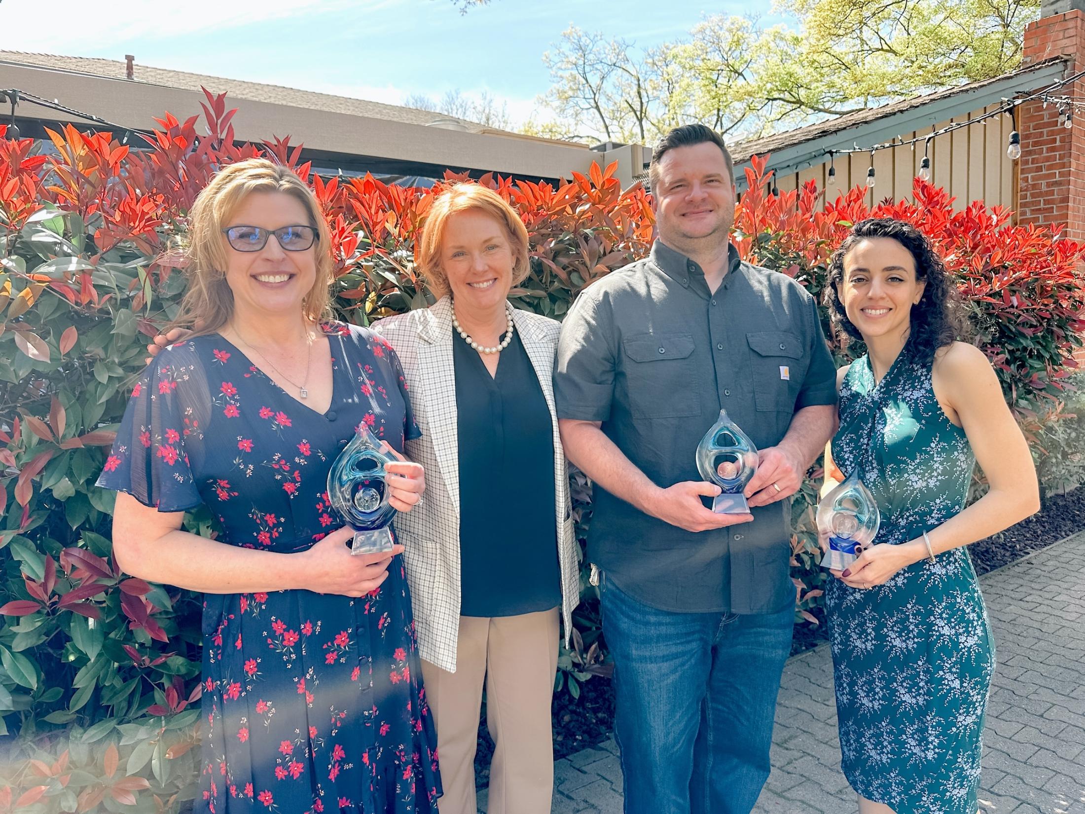Jennifer Thompson, Aaron Knapek, and Amber Sharpe stand holding awards, posing with Clare Shinnerl