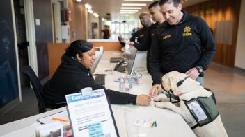 Yellow Labrador retriever stands on its hind legs against a welcome desk. The person behind the desk pets the dog while the handler, a CORE officer, stands on the other side