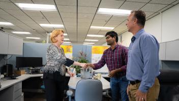 Woman shakes hands with two men outside of a cubicle