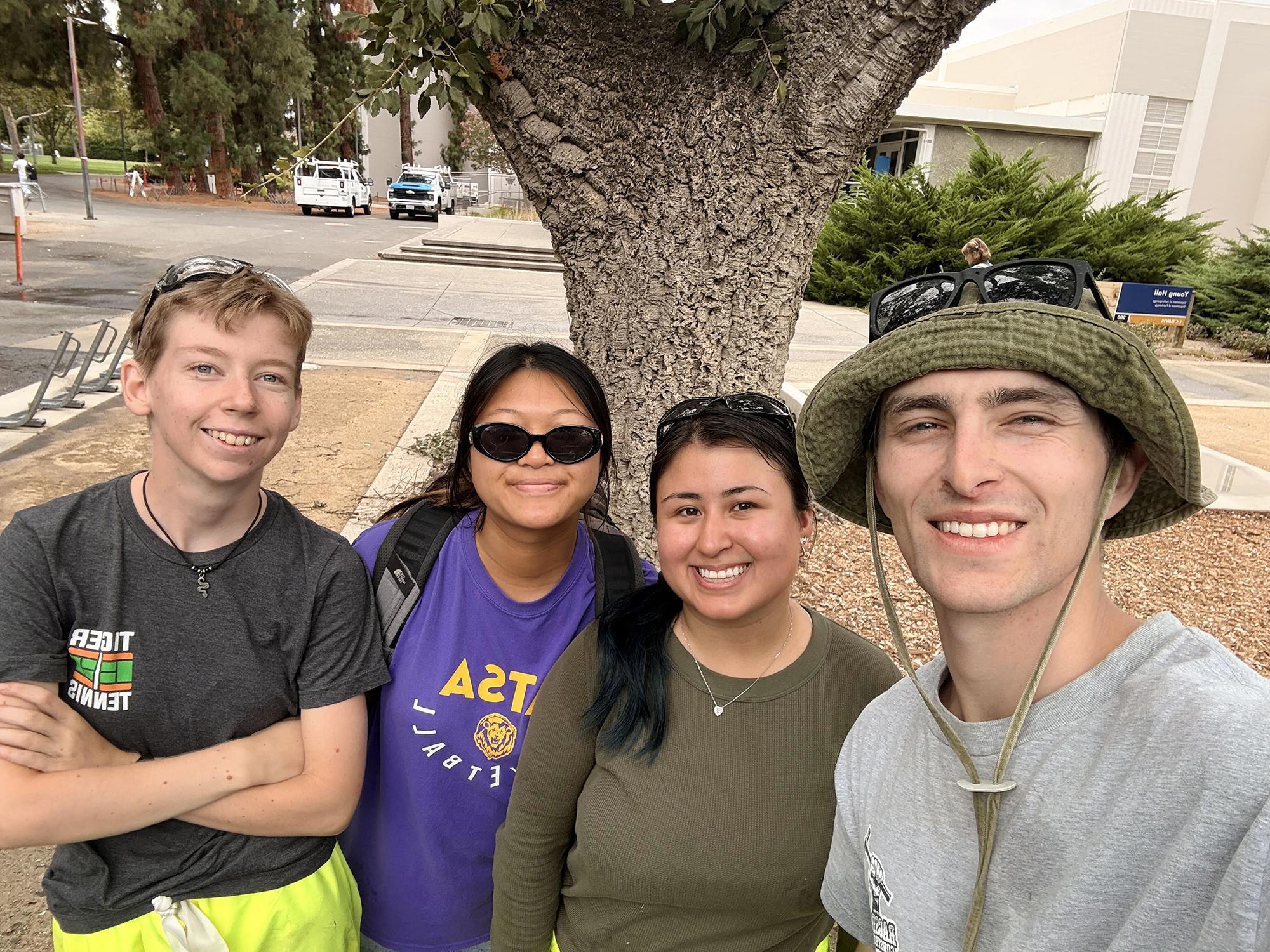 Four smiling students in a group selfie by a tree.