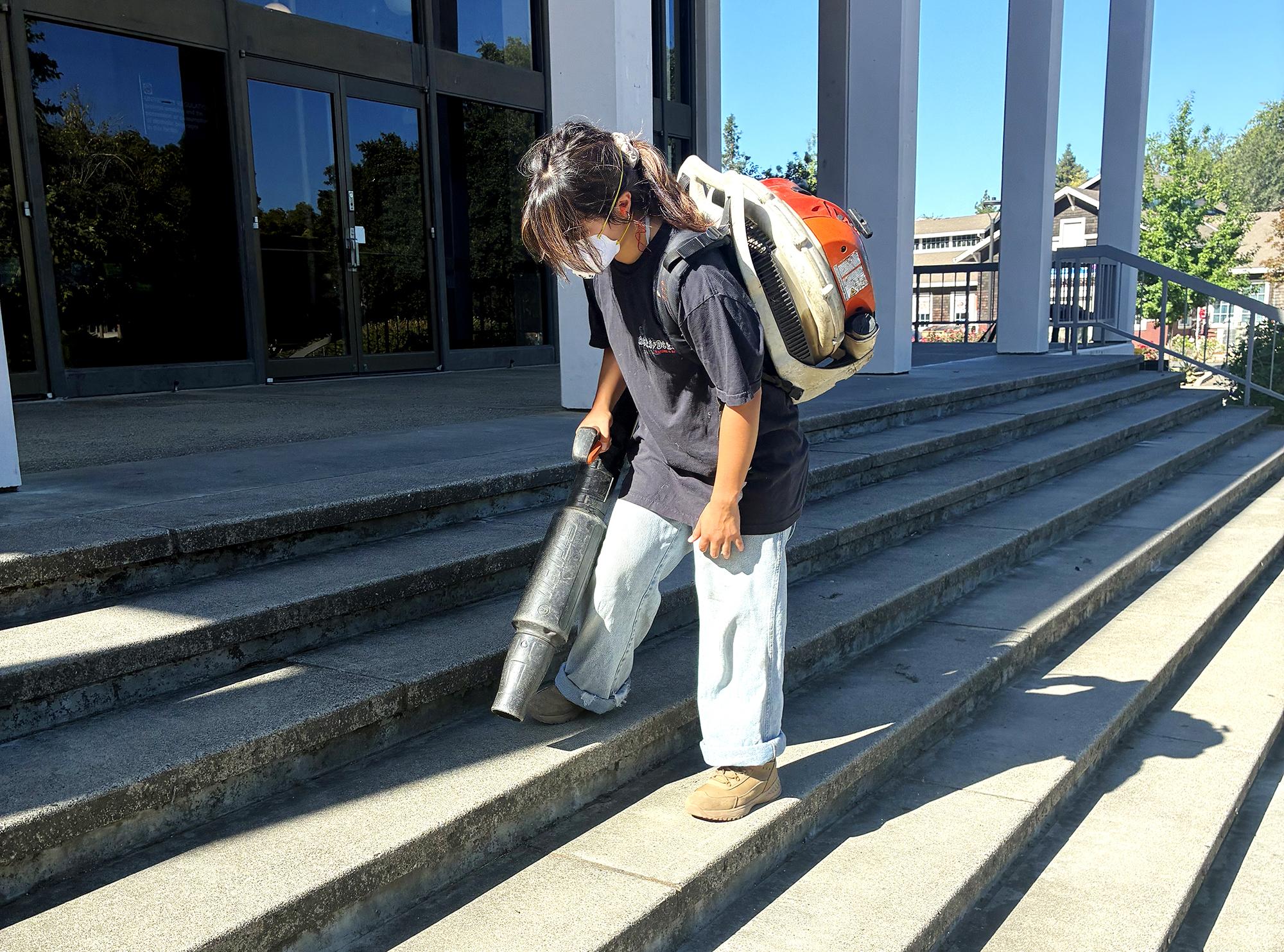 Student with a dust mask using a leaf blower on the front steps of Peter A Rock Hall.