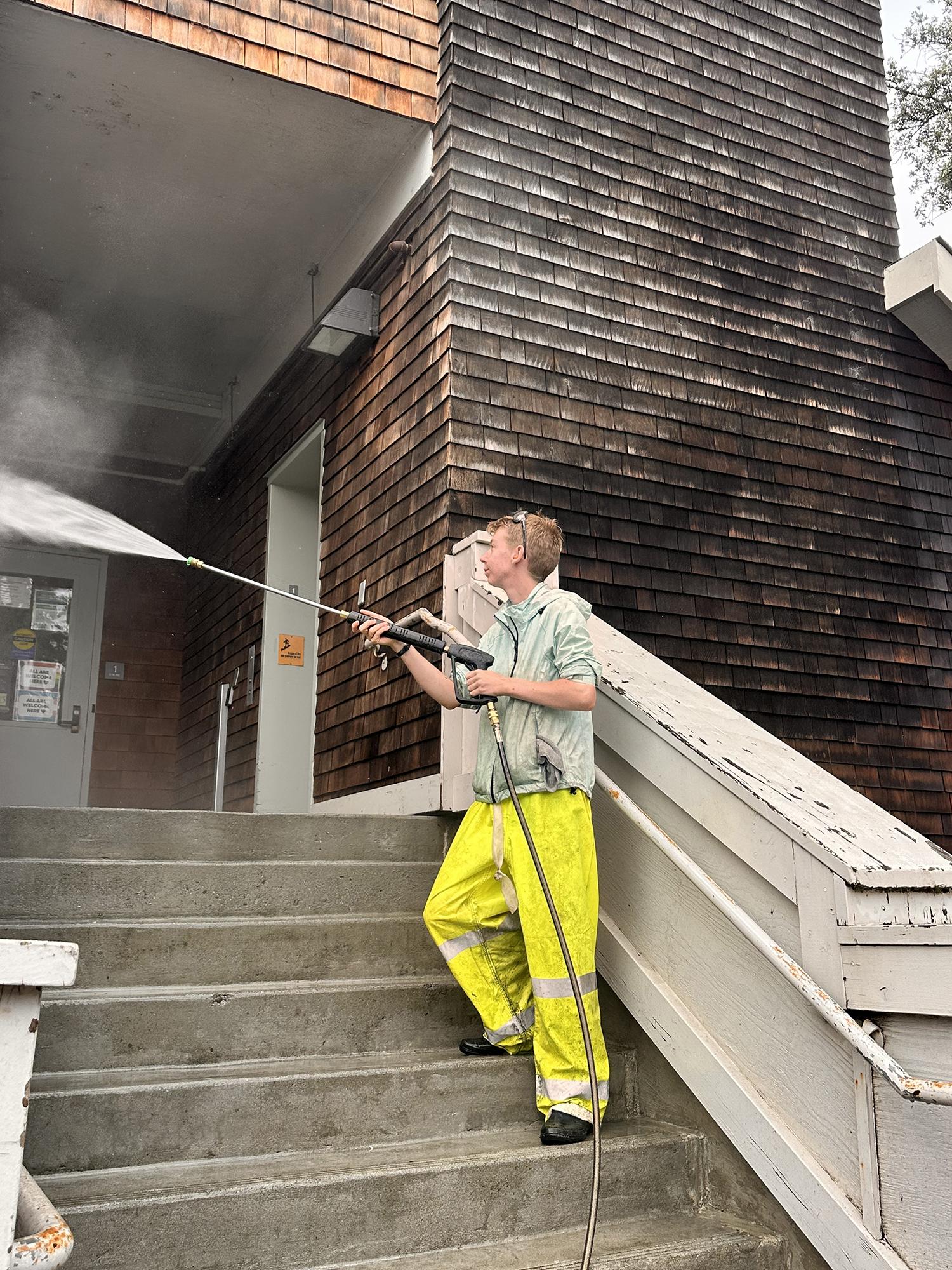 Student in yellow hi-vis pants pressure-washing a wooden building.