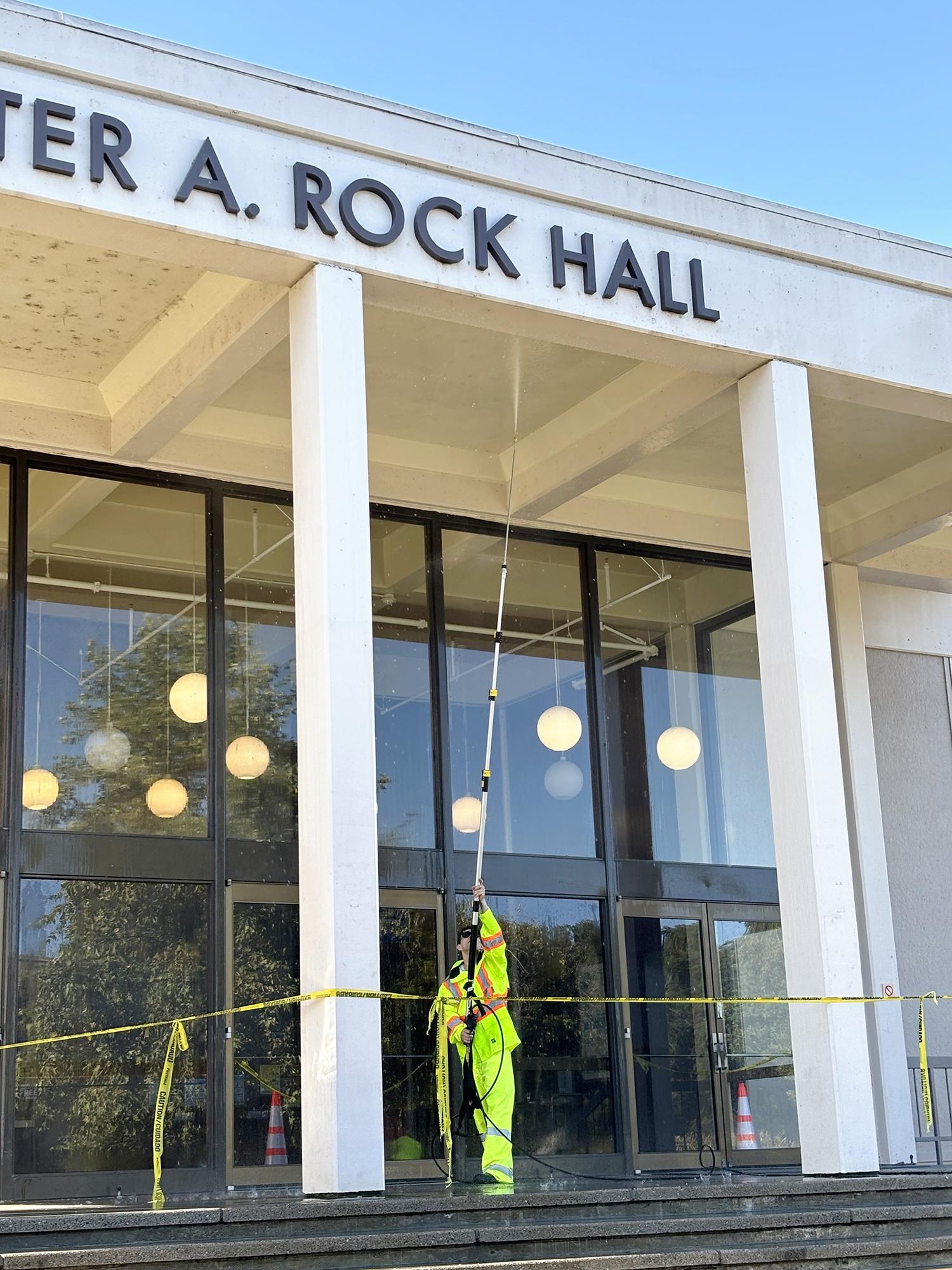 Student in a neon yellow safety suit behind caution tape pressure washing the awning of Peter A Rock Hall.