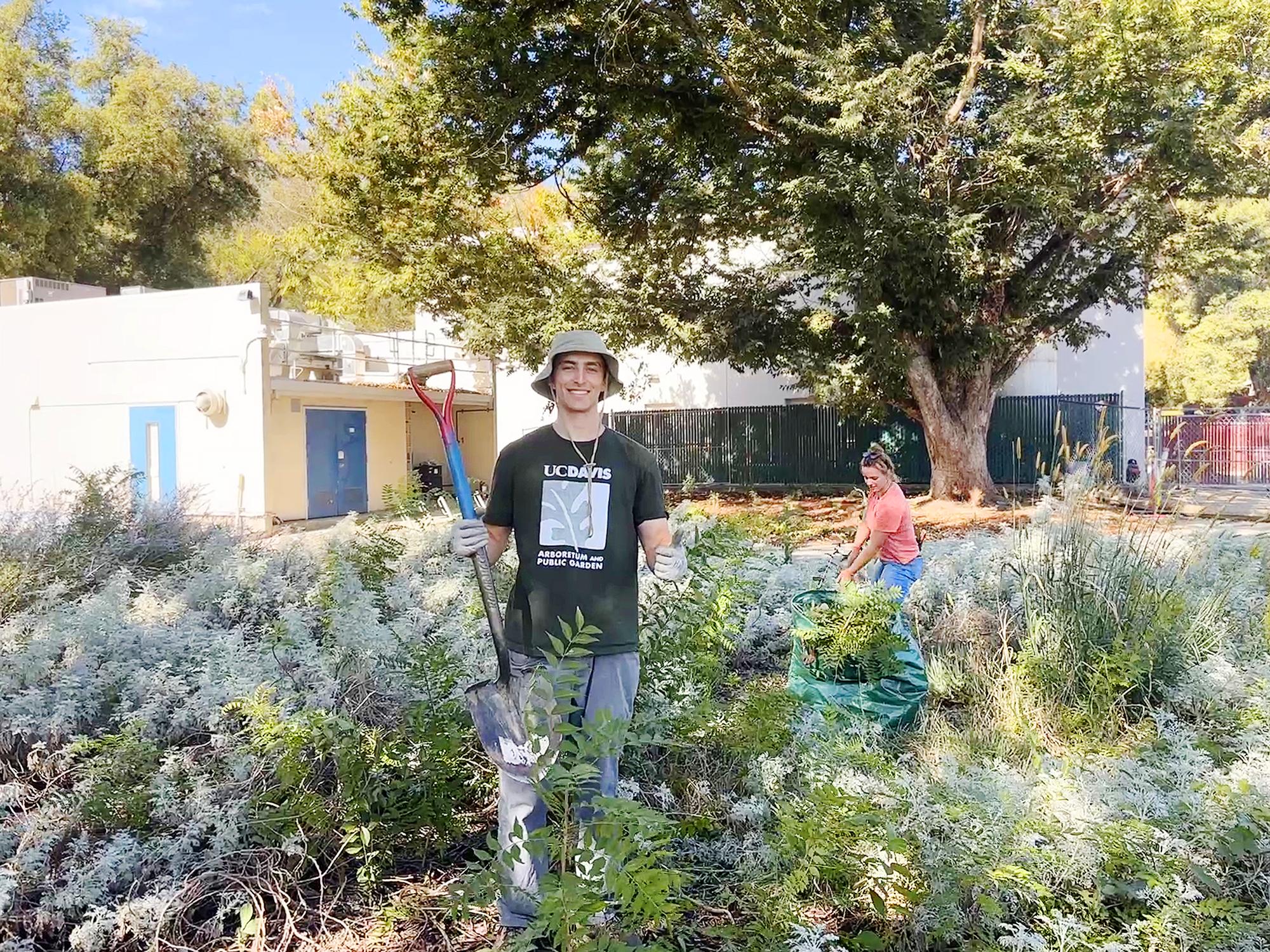 Student in an Arboretum bucket hat posing with a shovel and giving a thumbs up in a lush green area.