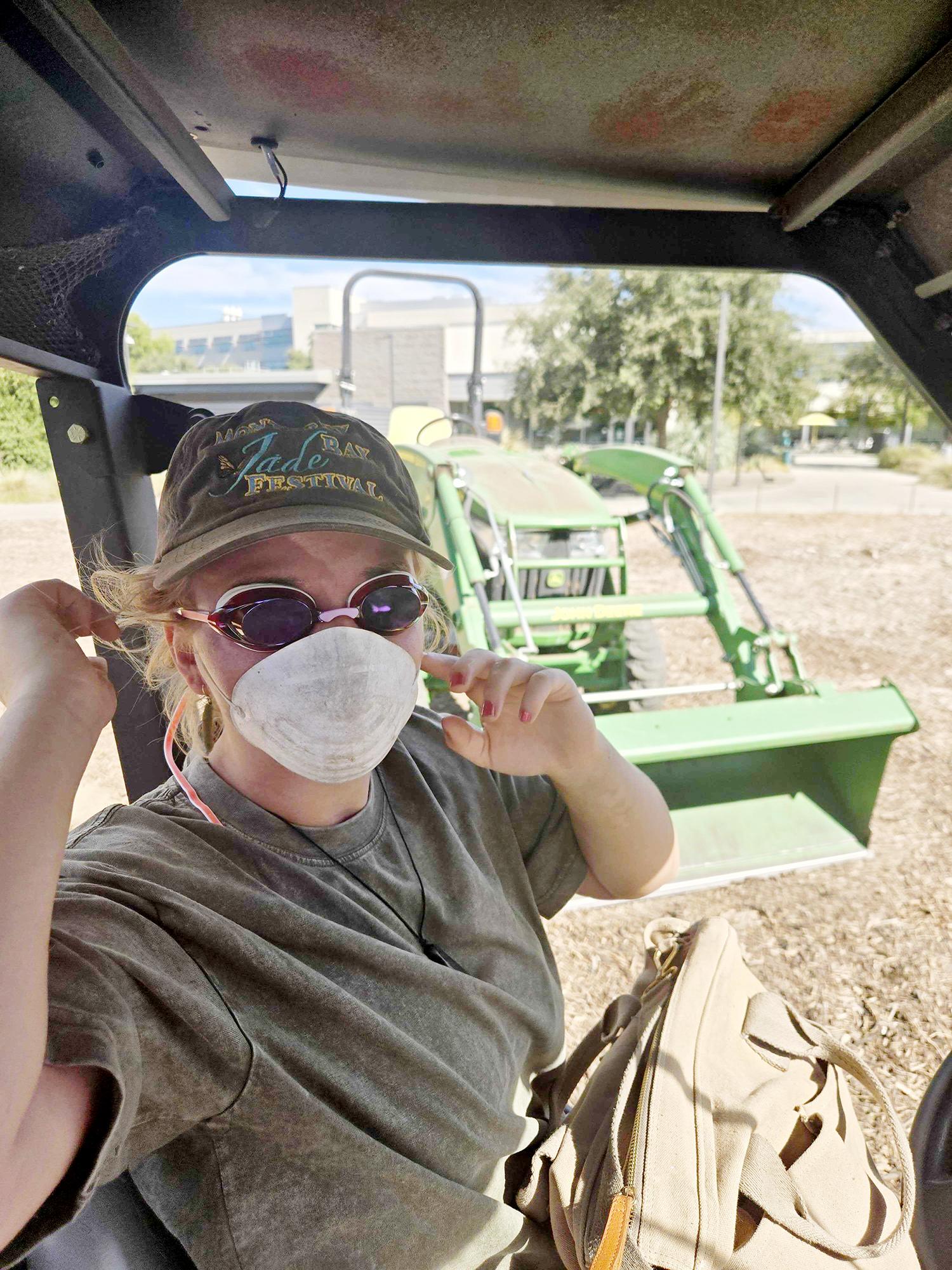 Student wearing a white dust mask inside tractor cab with another green tractor in the background.