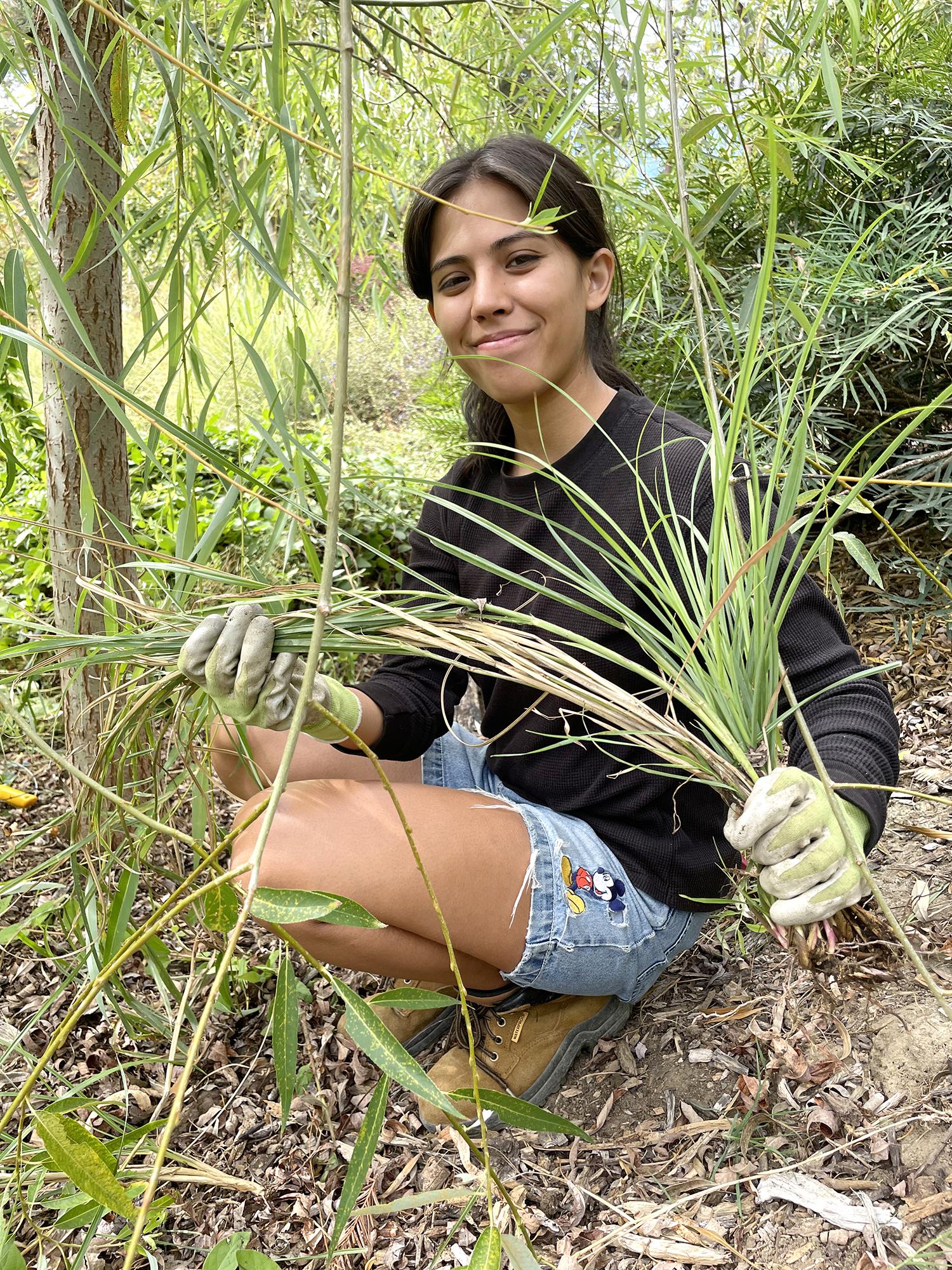 Student kneeling outdoors holding up a clumps of weeds and looking proudly at the camera.