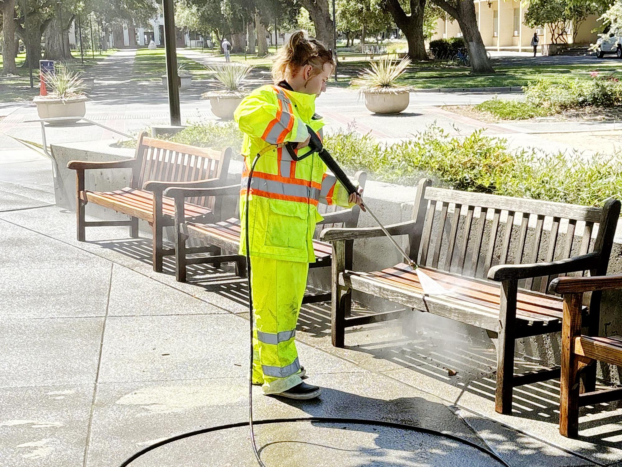 Student in yellow hi-vis suit pressure washing a bench.