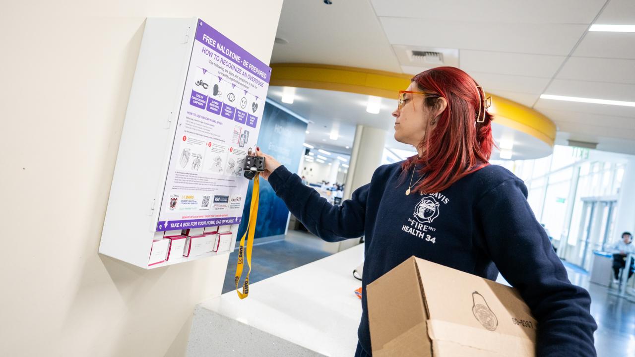 An EMT in a UC Davis shirt with a box of supplies, unlocks the Narcan cabinet in the Memorial Union