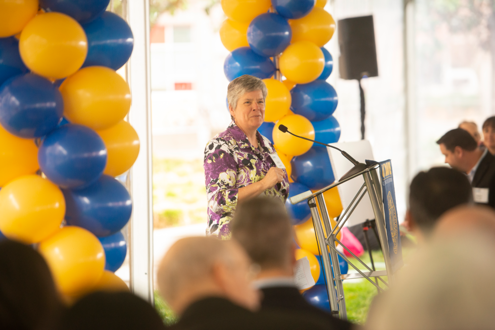 Kelly Ratliff speaking from a podium, surrounded by blue and gold balloons