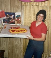 Kelly Ratliff holding a large pizza box, with 1980s era decor