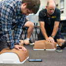Two men kneeling in a classroom practicing CPR on adult training mannequins