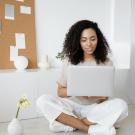 A woman sits cross legged on the floor while using a silver laptop.